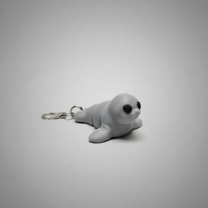 May include: A light gray seal keychain with black eyes and a silver keyring. The seal is lying down, with its flippers extended. The keychain is a cute, whimsical accessory.