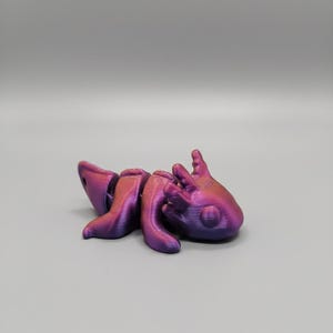 Puede incluir: Figura de ajolote impresa en 3D en tonos morados y rosas. La criatura, similar a una salamandra, tiene una cabeza detallada, cuatro patas y una cola. El ajolote está sobre una superficie gris.