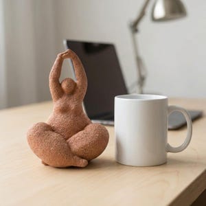 Könnte beinhalten: Eine terrakottafarbene Yoga-Figur mit erhobenen Armen, im Schneidersitz. Rechts befindet sich eine weiße Keramiktasse. Die Figur steht auf einer hellen Holzoberfläche, im Hintergrund ein Laptop und eine Schreibtischlampe.
