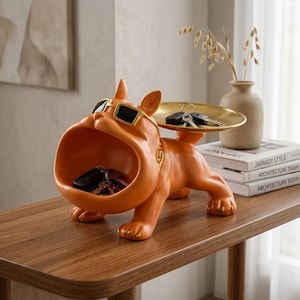 Peut inclure: Figurine de bouledogue orange avec des lunettes de soleil, une grande gueule ouverte et un plateau doré. Le bouledogue tient des clés dans sa bouche et sur le plateau. La figurine est sur une table en bois.