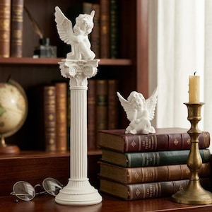 Puede incluir: Dos estatuas de querubines blancos, una sobre una columna estriada y la otra sobre libros. Los querubines tienen alas detalladas y son de estilo clásico. También hay un candelabro de latón con una vela encendida.