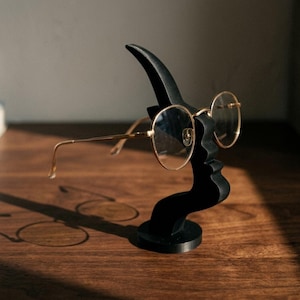 Peut inclure: Un présentoir à lunettes noir en forme de visage, tenant une paire de lunettes rondes à monture dorée. Le présentoir est posé sur une surface en bois, avec les lunettes projetant une ombre.