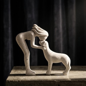 May include: A speckled white sculpture of a person leaning down to kiss a dog. The person's hair flows backward, and the dog stands on all fours. The sculpture is on a concrete surface against a dark background.