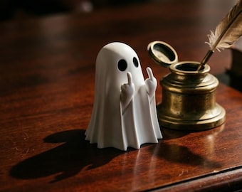 May include: A white ghost figurine with black eyes and raised middle fingers. The ghost is standing on a dark wooden surface next to a brass inkwell with a feather quill. The figurine has a minimalist design.