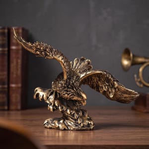 Peut inclure: Une figurine d'aigle aux tons bronze représentée dans une pose dynamique avec les ailes entièrement déployées et les serres tendues vers le bas. La sculpture présente des détails de plumes complexes et une finition métallique patinée.