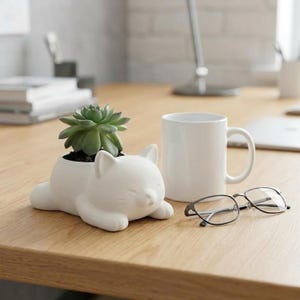 Puede incluir: Una maceta blanca con forma de gato que contiene una planta suculenta verde. Una taza blanca y un par de gafas están sobre un escritorio de madera. La maceta de gato tiene una cara sonriente.