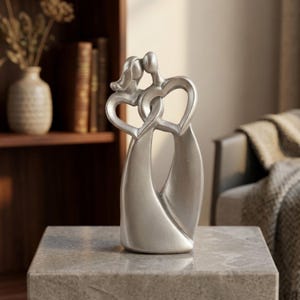 Puede incluir: Una escultura de color plateado que representa dos figuras abstractas de pie una al lado de la otra. La parte superior de los cuerpos está curvada para formar dos corazones entrelazados, mientras que la parte inferior se une en una base sólida.