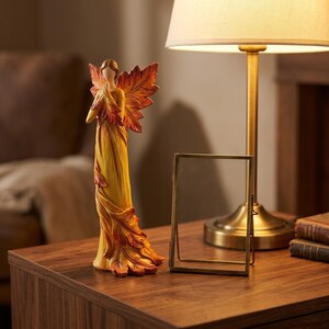 Peut inclure: Une figurine décorative d'ange avec des ailes en forme de feuilles et une robe fluide dans les tons de jaune, d'orange et de rouge. La figurine est sur une surface en bois, à côté d'une lampe dorée et d'un petit cadre photo.