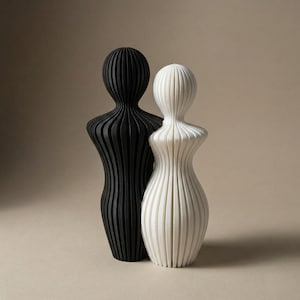 May include: Two abstract sculptures, one black and one white, stand side-by-side. Each figure has a rounded head and a curvaceous body, with vertical lines creating a ribbed texture. The sculptures are set against a neutral background.