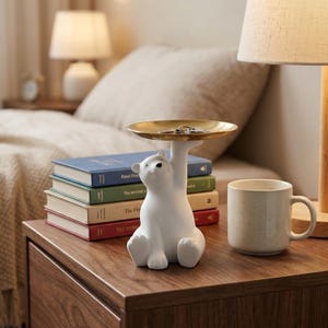 Puede incluir: Una figura de oso polar blanco sosteniendo una pequeña bandeja redonda dorada. La bandeja contiene monedas. El oso está sobre una mesita de noche de madera junto a una pila de libros y una taza blanca moteada.
