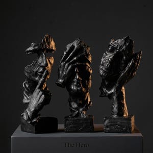 Peut inclure: Trois sculptures noires de figures humaines dans différentes poses, chacune sur une base carrée. Les sculptures ont un aspect texturé et rugueux et sont exposées sur une surface gris foncé. Le texte "The Hero" est visible.