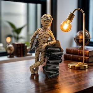 Peut inclure: Une figurine fantaisiste de momie portant des lunettes, appuyée contre une pile de livres noirs. La figurine est beige avec des rayures noires. Une lampe de bureau dorée avec une ampoule allumée et un globe sont également visibles.