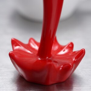 May include: Close-up of a glossy red decorative object. The object has a flower-like base with pointed petals and a long, cylindrical stem. The surface is smooth and reflects light.