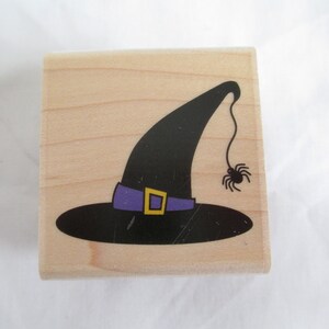 May include: Wooden stamp featuring a black witch hat with a purple band and gold buckle. A black spider hangs from the hat. The stamp is perfect for Halloween crafts and decorations.