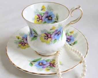 May include: A white teacup and saucer with a floral pattern of purple, yellow, and blue pansies. A pearl necklace is draped over the saucer.