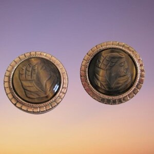 Puede incluir: Un par de pendientes de color dorado con piedras redondas de ojo de tigre. Las piedras tienen un diseño tallado de una cara.