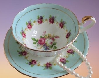 May include: A light blue teacup and saucer with a floral pattern and gold trim. A pearl necklace is draped around the saucer.