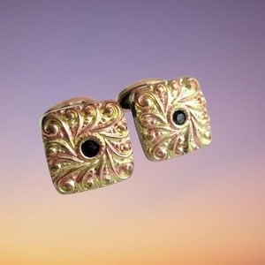 May include: A pair of gold cufflinks with a square design. Each cufflink has a black stone in the center and intricate floral patterns.