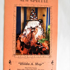 Puede incluir: Una tarjeta naranja con el texto "SEW SPECIAL" y una imagen enmarcada de una muñeca de bruja hecha a mano. La muñeca tiene una cara blanca, un sombrero negro y naranja y un cuerpo similar a una fregona naranja y negra. El texto "Hilda A. Mop" está debajo.