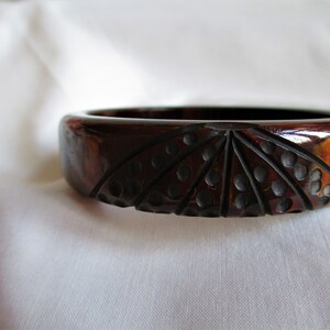 May include: A dark brown carved plastic bangle bracelet with a geometric design. The design features a fan-like pattern with black dots.