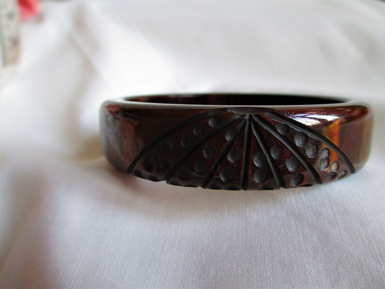 May include: A dark brown carved plastic bangle bracelet with a geometric design. The design features a fan-like pattern with black dots.