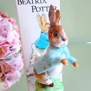 Puede incluir: Una figura de cerámica de Peter Rabbit, con una chaqueta azul, de pie sobre una base verde. La figura está frente a una caja con las palabras "BEATRIX POTTER" impresas. Un ramo de flores rosas está en el fondo.