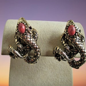 May include: A pair of gold-tone earrings featuring a detailed lizard design with a pink stone accent.
