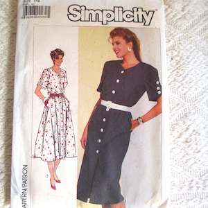 Pode incluir: Um envelope de molde de costura Simplicity, número 7889, tamanho 14. A frente exibe ilustrações de dois estilos de vestido: um em uma cor escura sólida com cinto e botões brancos, e outro em branco com bolinhas vermelhas e cinto vermelho.