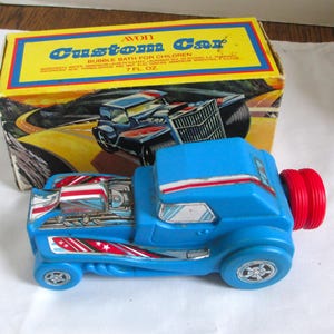 Puede incluir: Un coche de juguete de plástico azul con detalles rojos y blancos, y una tapa roja en la parte trasera. El coche está delante de una caja amarilla con las palabras "Custom Car" y una ilustración de un coche.