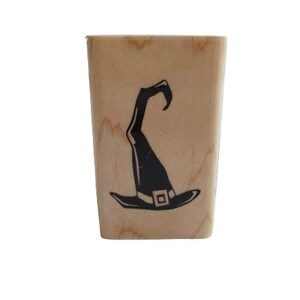 May include: A wooden stamp with a black witch's hat design. The hat has a wide brim, a pointed crown, and a buckle detail. The stamp is rectangular and made of light-colored wood, perfect for Halloween crafts.