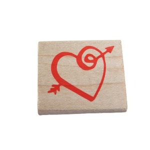 May include: A small, square wooden stamp with a red heart pierced by an arrow. The heart and arrow design is centered on the light-colored wood block. The arrow's point is on the left, and the fletching is on the lower left.