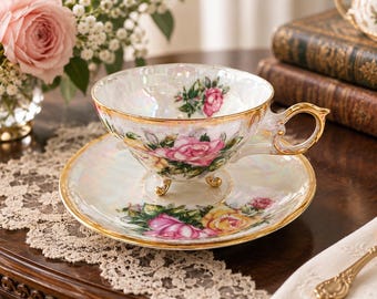 Puede incluir: Un juego de taza de té y platillo de porcelana con acabado iridiscente, bordes dorados y un estampado floral de rosas rosas y amarillas. La taza se apoya sobre tres pequeñas patas doradas e incluye un asa decorativa dorada.