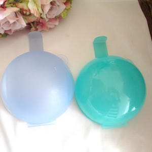Puede incluir: Dos recipientes esféricos translúcidos, uno azul claro y otro turquesa, cada uno con una extensión similar a un asa. Los recipientes están sobre una superficie blanca, con un arreglo floral en el fondo.
