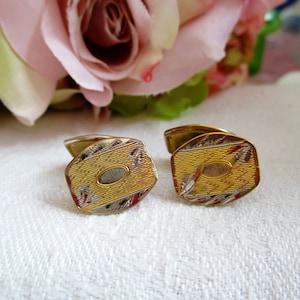 Puede incluir: Un par de gemelos de oro con un diseño geométrico. Los gemelos tienen un borde plateado y un centro dorado con un patrón texturizado.