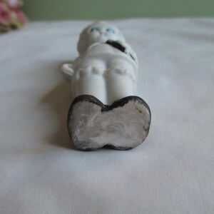 May include: A small, white ceramic figurine of a child with blue eyes and black shoes. The figurine is lying on its back, with its arms at its sides. The figurine has a black bow tie and a yellow accent on the back.