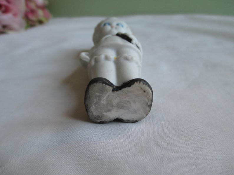 May include: A small, white ceramic figurine of a child with blue eyes and black shoes. The figurine is lying on its back, with its arms at its sides. The figurine has a black bow tie and a yellow accent on the back.