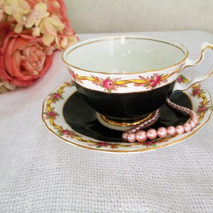 May include: A black and white teacup and saucer with a gold trim and pink floral design. A pink pearl necklace is draped over the saucer.