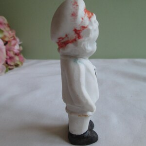 May include: A small, vintage porcelain figurine of a child in a white outfit with a matching hat. The hat has red accents. The figurine has black shoes and a small black detail on the chest. The figurine is standing.
