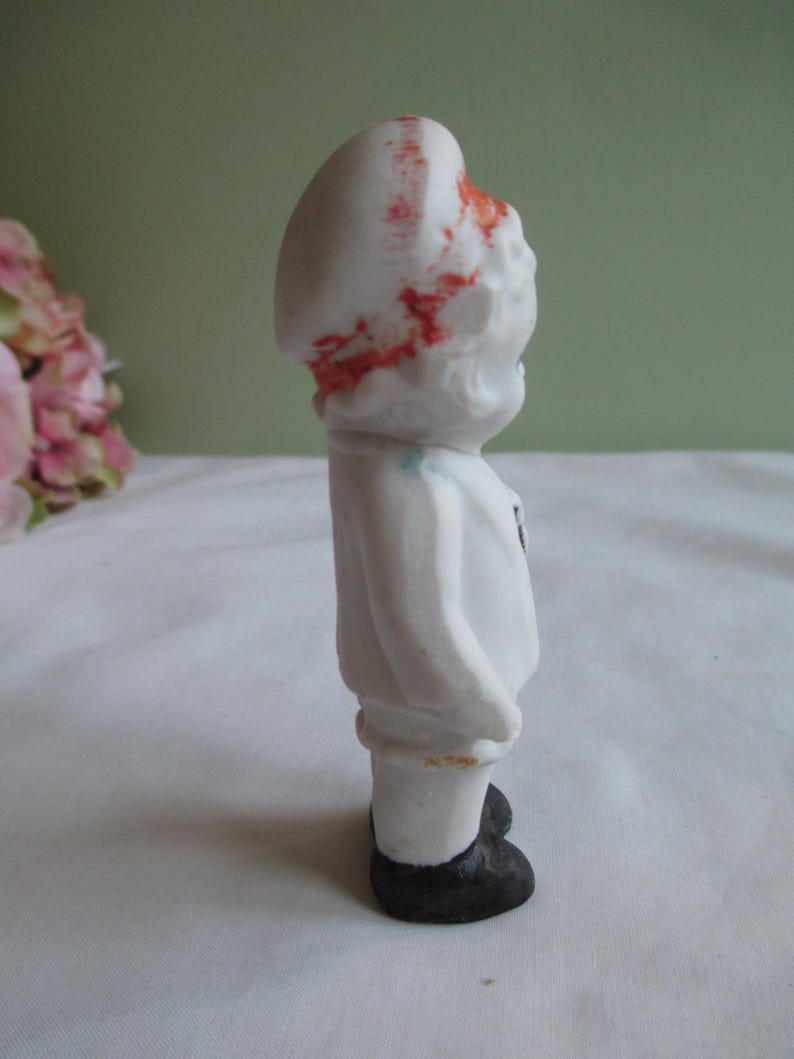 May include: A small, vintage porcelain figurine of a child in a white outfit with a matching hat. The hat has red accents. The figurine has black shoes and a small black detail on the chest. The figurine is standing.