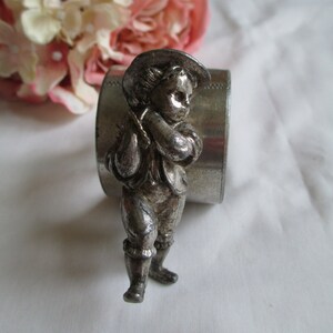 May include: A silver-toned napkin ring featuring a detailed figurine of a young boy in a hat and period clothing. The ring is cylindrical with a textured edge, and the boy is holding a musical instrument. The background includes pink flowers.