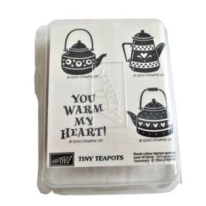 May include: A set of three black and white rubber stamps featuring teapots with heart designs. The stamps are packaged in a clear plastic case with the text "You warm my heart!" and "Tiny Teapots".