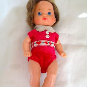 May include: A vintage baby doll with blue eyes, rosy cheeks, and light brown hair. The doll is dressed in a red onesie with a white collar and a decorative band with red and green floral designs. The doll is made of plastic and is a classic toy.