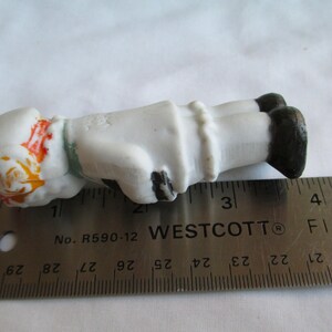 May include: A small, porcelain figurine of a person lying down, with a white body, black shoes, and a white hat with orange and yellow accents. The figurine is approximately 3 inches long, as measured by a ruler.
