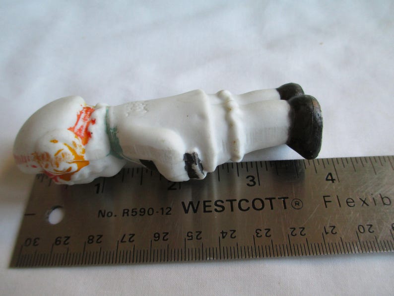May include: A small, porcelain figurine of a person lying down, with a white body, black shoes, and a white hat with orange and yellow accents. The figurine is approximately 3 inches long, as measured by a ruler.