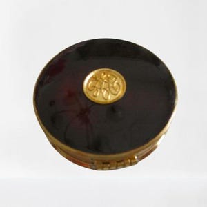 May include: A round, dark brown compact with a gold-colored rim and a gold emblem on the top. The compact appears to be made of a shiny material, reflecting light. The emblem is a circular design with ornate detailing.