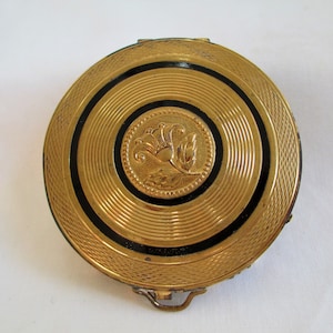 May include: A gold compact with a black enamel design and a floral motif in the center. The compact has a hinged lid and a decorative clasp.