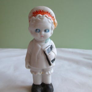 May include: A small, vintage porcelain doll with blue eyes, red lips, and a white outfit. The doll wears a white hat with orange trim and holds a black and white object. The doll's shoes are black.