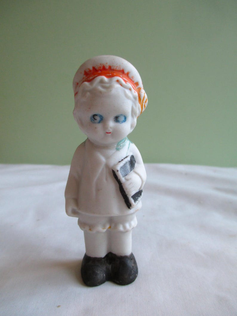 May include: A small, vintage porcelain doll with blue eyes, red lips, and a white outfit. The doll wears a white hat with orange trim and holds a black and white object. The doll's shoes are black.
