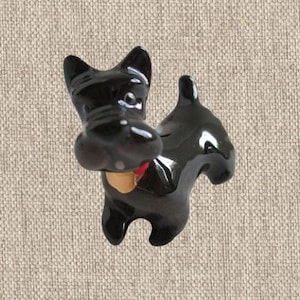 Op de afbeelding: Een glanzend zwart keramieken beeldje van een Scottish Terrier hond. De hond heeft puntige oren, een korte omhoogstaande staart en een kleine rode halsband met een beige penning om de nek. Het oppervlak van het beeldje is glad en reflecterend.