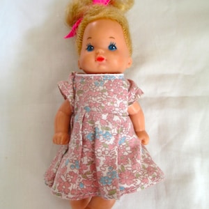 May include: A vintage doll with blonde hair in a bun, decorated with a pink bow. The doll has blue eyes and wears a pink floral dress. A classic toy.